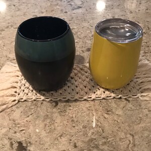 May include: Two insulated tumblers, one black with a teal band and one yellow, on a white crocheted coaster with fringe.