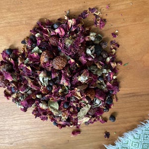 Op de afbeelding: Een close-up van een stapel gedroogde rozenblaadjes, jeneverbessen en andere botanicals. Het mengsel is diep rood en paars, met wat bruine en groene accenten.