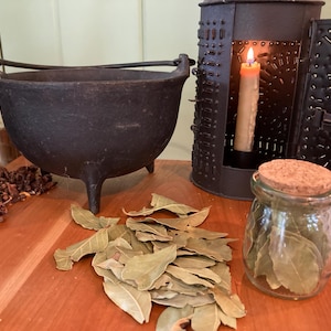 Puede incluir: Un caldero de hierro fundido negro con tres patas, una linterna de velas y un tarro de vidrio con tapa de corcho. Hojas de laurel secas y especias dispuestas sobre una superficie de madera.