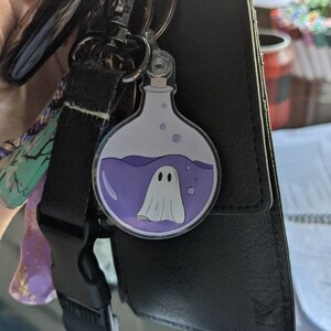 May include: A keychain with a round, clear charm featuring a cartoon ghost inside a potion bottle. The bottle has purple liquid and bubbles. The keychain is attached to a black strap and a black wallet.