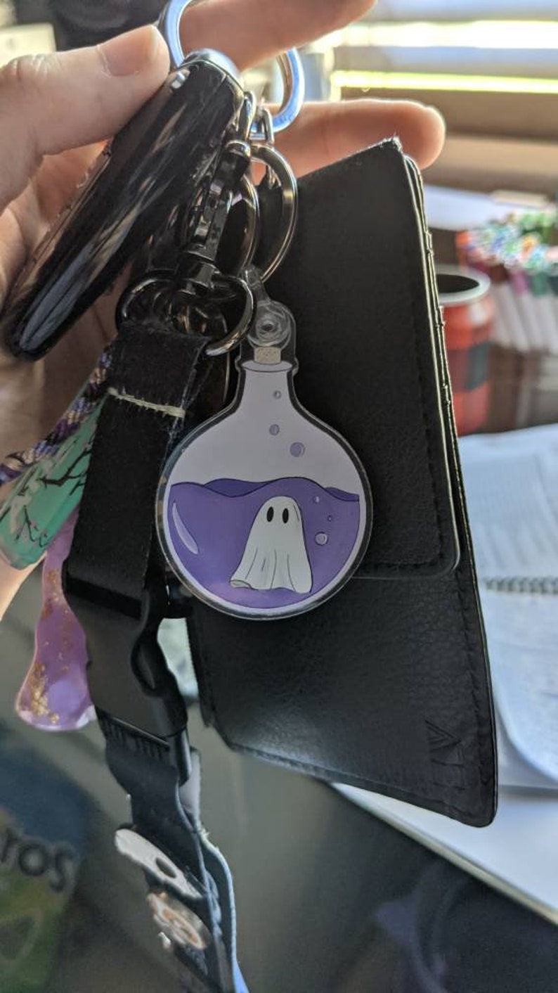 May include: A keychain with a round, clear charm featuring a cartoon ghost inside a potion bottle. The bottle has purple liquid and bubbles. The keychain is attached to a black strap and a black wallet.