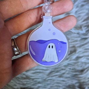 May include: A clear acrylic keychain featuring a cartoon ghost inside a purple potion bottle. The bottle has a white top and a silver keyring. The ghost has a white body and two black eyes. The keychain is held by a hand.