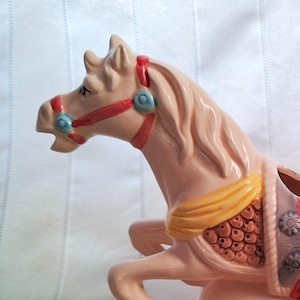 May include: A ceramic carousel horse creamer in a soft pink hue. The horse features a red bridle, a yellow saddle blanket, and a decorative saddle. The handle is shaped like a horse's tail. A charming addition to any kitchen.