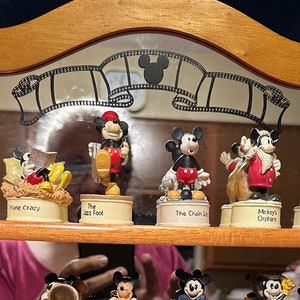 May include: A wooden shelf unit with three shelves displays a collection of 24 miniature figurines of Mickey Mouse in various poses and settings. Each figurine is in a small glass dome with a label underneath. The figurines are from the Disney Traditions collection.