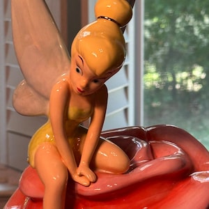 May include: A ceramic figurine of a fairy with blonde hair and yellow attire, seated on a large, pink rose. The fairy has large, white wings and is posed with her hands clasped. The rose has green leaves at the base.