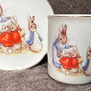 May include: A white ceramic teacup and saucer set with a gold rim. The set features a colorful illustration of Peter Rabbit and his siblings.