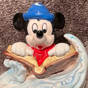 Peut inclure: Une figurine en c&eacute;ramique de Mickey Mouse portant un chapeau bleu avec une lune croissante, lisant un livre assis dans une vague. La figurine est peinte dans des tons de bleu, blanc, rouge et or.