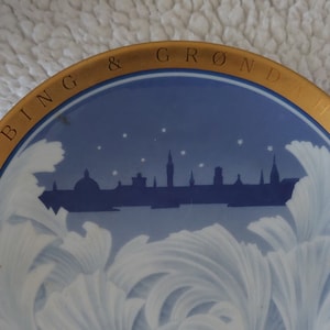May include: A decorative plate with a blue and white design featuring a city skyline and stylized clouds. The plate has a gold rim and the text "BING & GRØNDAHL 1995 AR".