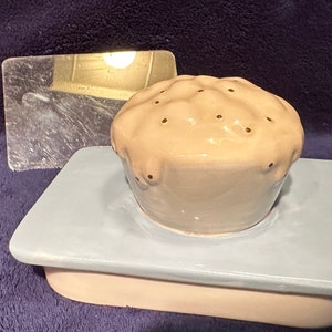 May include: A ceramic cupcake shaped dish with a light blue base and a light brown top with brown dots. The dish is sitting on a light pink base.