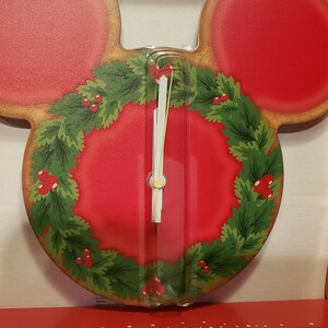 May include: A Mickey Mouse-shaped wall clock with a red face and ears. The clock face features a green wreath with red berries. The clock is in a red and white box with a festive design.