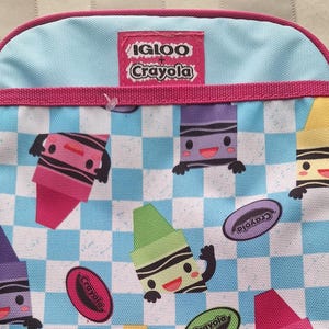 May include: A rectangular Igloo Crayola lunch bag. The bag features a blue and white checkered pattern with cartoon crayons in various colors. The bag has a pink trim and a blue top with the Igloo Crayola logo.