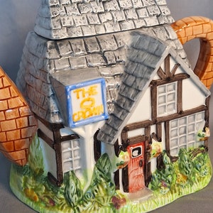 May include: A ceramic teapot shaped like a cottage with a gray tiled roof, brown brick chimney, and a sign that reads "The Crown". The teapot has a brown brick spout and a brown handle.