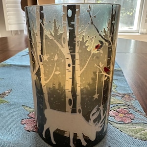 May include: A cylindrical glass candle holder with a winter scene design. The design features white birch trees, a deer silhouette, and red birds. The background has a gradient of blue and green. The candle holder is sitting on a blue table runner.