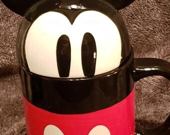 May include: A black and red ceramic mug with a Mickey Mouse design. The mug has a lid with Mickey Mouse ears.