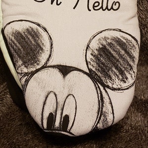 May include: A pair of oversized oven mitts with a light blue background and a black and white sketch of Mickey Mouse's face. The text "Oh Hello" is written in black cursive above Mickey's face. The packaging shows a red and white Mickey Mouse with the text "Disney Mickey Mouse" and "2PK Oversized Mini Mitts".