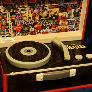 May include: A black and red ceramic record player with a Beatles logo and a record on the turntable. The lid is open and features a collage of Beatles images.