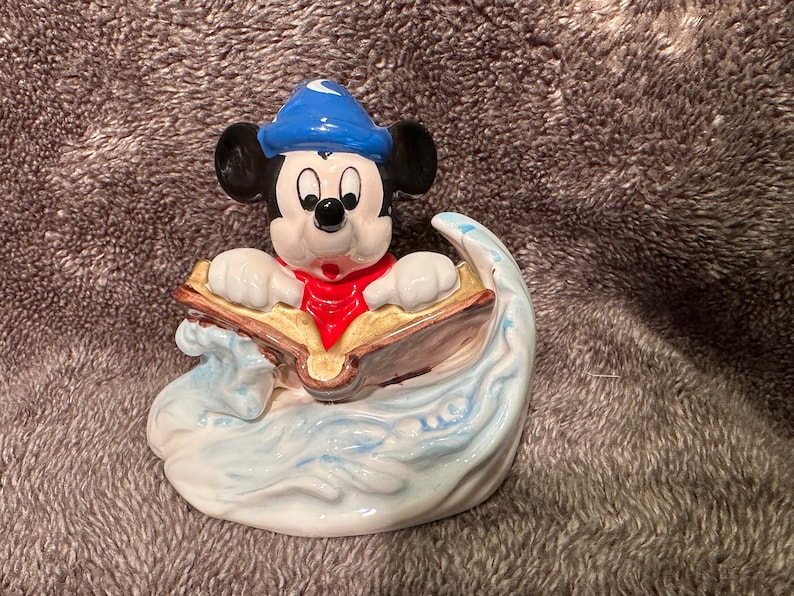 Peut inclure: Une figurine en c&eacute;ramique de Mickey Mouse portant un chapeau bleu avec une lune croissante, lisant un livre assis dans une vague. La figurine est peinte dans des tons de bleu, blanc, rouge et or.