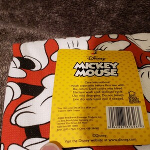 May include: A yellow and red care label for a Mickey Mouse kitchen towel. The label has care instructions for washing and ironing the towel. The label also includes the Disney logo and website address.