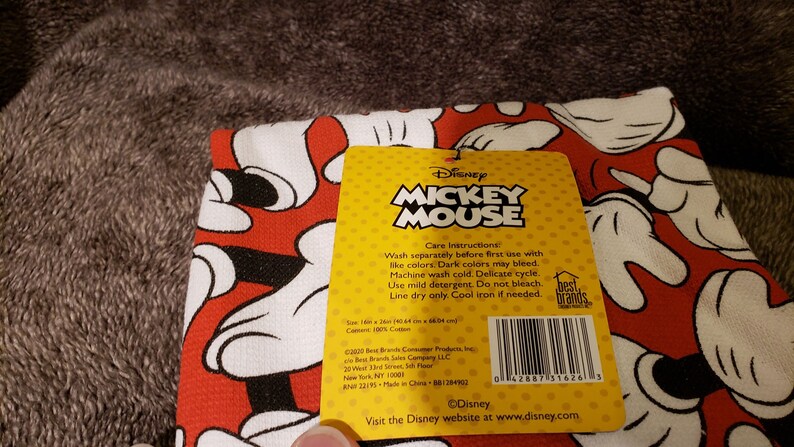 May include: A yellow and red care label for a Mickey Mouse kitchen towel. The label has care instructions for washing and ironing the towel. The label also includes the Disney logo and website address.