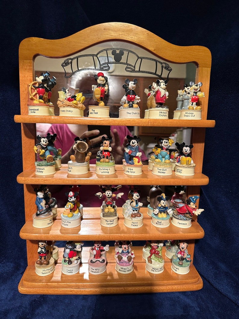 May include: A wooden shelf unit with three shelves displays a collection of 24 miniature figurines of Mickey Mouse in various poses and settings. Each figurine is in a small glass dome with a label underneath. The figurines are from the Disney Traditions collection.