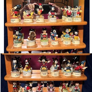 May include: A wooden shelf unit with a mirror on top displays a collection of 24 miniature figurines of Mickey Mouse in various poses. Each figurine is on a small, round base with a different scene or theme depicted on it. The scenes include Mickey Mouse in a boat, playing music, building a house, and more. The shelf unit is made of light brown wood and has a dark brown stain.