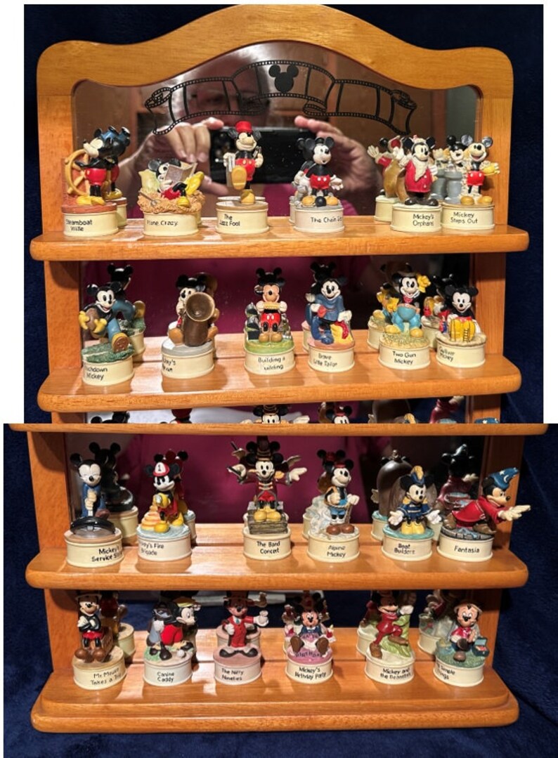 May include: A wooden shelf unit with a mirror on top displays a collection of 24 miniature figurines of Mickey Mouse in various poses. Each figurine is on a small, round base with a different scene or theme depicted on it. The scenes include Mickey Mouse in a boat, playing music, building a house, and more. The shelf unit is made of light brown wood and has a dark brown stain.