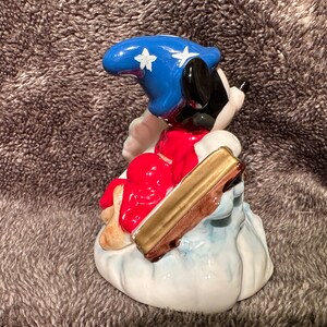 Peut inclure: Une figurine en c&eacute;ramique de Mickey Mouse habill&eacute; en sorcier, portant un chapeau bleu avec des &eacute;toiles blanches et une robe rouge. Il est assis sur un nuage et tient un b&acirc;ton dor&eacute;.