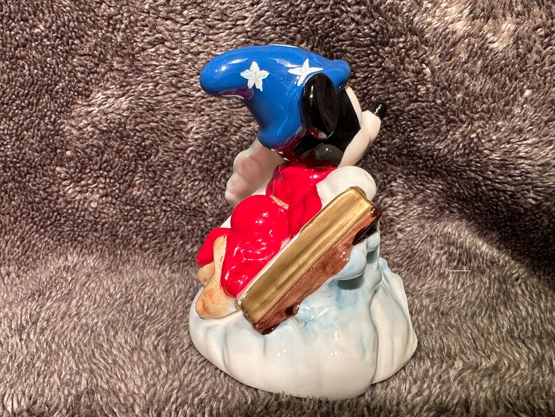 Peut inclure: Une figurine en c&eacute;ramique de Mickey Mouse habill&eacute; en sorcier, portant un chapeau bleu avec des &eacute;toiles blanches et une robe rouge. Il est assis sur un nuage et tient un b&acirc;ton dor&eacute;.