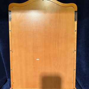 May include: The back of a wooden wall-mounted mirror with a scalloped top. The mirror has a light wood finish and is secured to the wall with two metal brackets.