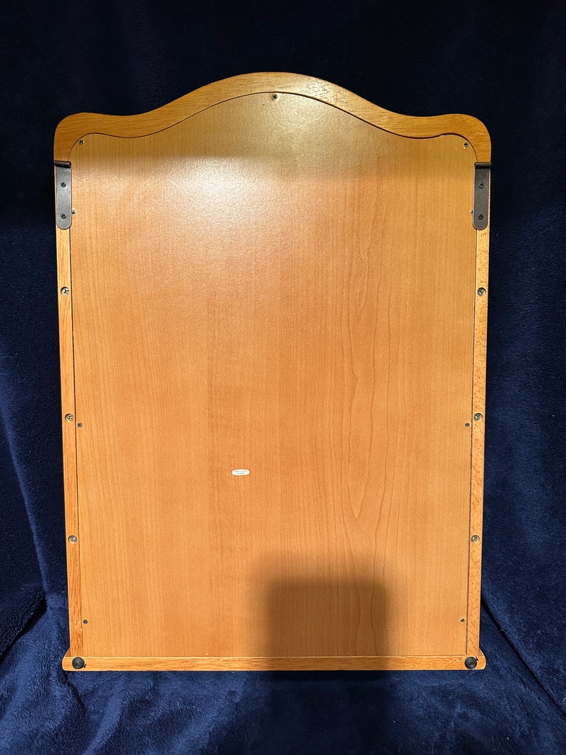 May include: The back of a wooden wall-mounted mirror with a scalloped top. The mirror has a light wood finish and is secured to the wall with two metal brackets.