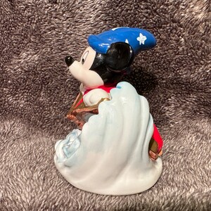 Peut inclure: Une figurine en c&eacute;ramique de Mickey Mouse habill&eacute; en sorcier. Il porte un chapeau bleu avec des &eacute;toiles blanches et une robe rouge. Il se tient debout sur une vague blanche.