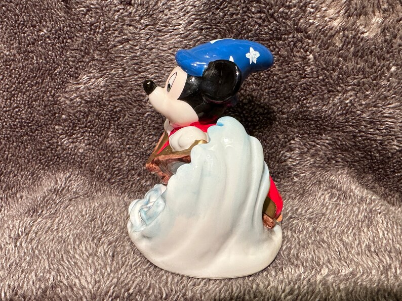 Peut inclure: Une figurine en c&eacute;ramique de Mickey Mouse habill&eacute; en sorcier. Il porte un chapeau bleu avec des &eacute;toiles blanches et une robe rouge. Il se tient debout sur une vague blanche.