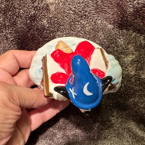 Peut inclure: Une figurine en c&eacute;ramique d'un chien noir et blanc portant un chapeau bleu avec une lune croissante et une &eacute;toile. Le chien est allong&eacute; sur le dos, les pattes en l'air, et un livre rouge et blanc sous la t&ecirc;te.
