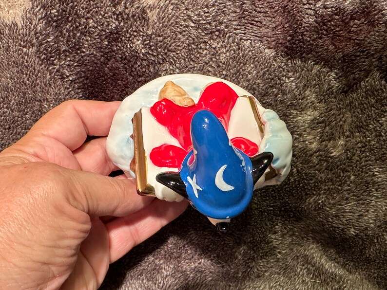 Peut inclure: Une figurine en c&eacute;ramique d'un chien noir et blanc portant un chapeau bleu avec une lune croissante et une &eacute;toile. Le chien est allong&eacute; sur le dos, les pattes en l'air, et un livre rouge et blanc sous la t&ecirc;te.
