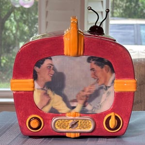 May include: A ceramic television-shaped decorative object in deep red and bright orange. The screen displays a vintage-style image of a person in a yellow sweater and a person in a white shirt interacting. It features two knobs, a speaker grille, and two antennae.