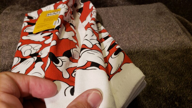 May include: A folded white and red kitchen towel with a Mickey Mouse hand print pattern. The towel has a yellow tag with the text "Mickey Mouse" on it.