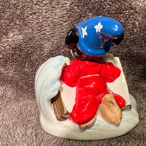 Peut inclure: Une figurine en c&eacute;ramique de Mickey Mouse habill&eacute; en sorcier, portant un chapeau bleu avec des &eacute;toiles blanches, une robe rouge et tenant un livre. Il se tient sur une vague blanche avec un livre dor&eacute;.