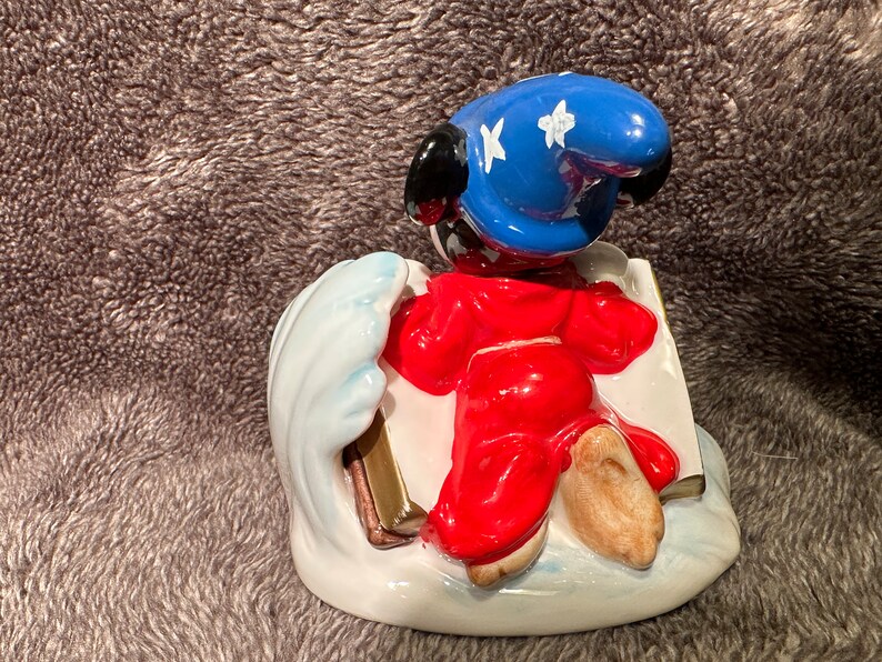 Peut inclure: Une figurine en c&eacute;ramique de Mickey Mouse habill&eacute; en sorcier, portant un chapeau bleu avec des &eacute;toiles blanches, une robe rouge et tenant un livre. Il se tient sur une vague blanche avec un livre dor&eacute;.