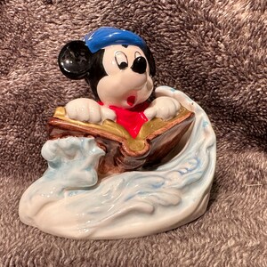Peut inclure: Une figurine en c&eacute;ramique de Mickey Mouse portant un chapeau de marin bleu et une &eacute;charpe rouge, tenant un livre tout en surfant sur une vague.