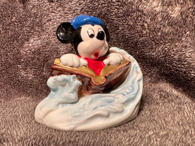 Peut inclure: Une figurine en c&eacute;ramique de Mickey Mouse portant un chapeau de marin bleu et une &eacute;charpe rouge, tenant un livre tout en surfant sur une vague.