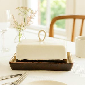 Op de afbeelding: Een witte rechthoekige botervloot met een lusvormige handgreep op een donkerbruine schaal. Een witte mok, bord en bestek liggen op een wit tafelkleed. Een kleine vaas met bloemen en een wijnglas staan ook op tafel.
