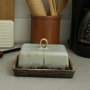 Puede incluir: Una mantequera de cerámica con un esmalte azul claro y marrón, con un asa de bucle. Utensilios de cocina de madera, incluyendo un rodillo, cucharas y un tenedor, están en una olla de cerámica marrón.