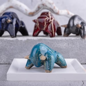 May include: Five ceramic bull figurines in various colors, including blue, red, and gray. The figurines are arranged on a white surface with a wooden branch in the background.