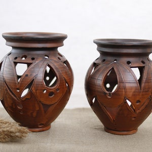 Puede incluir: Dos jarrones de cerámica marrón con intrincados diseños recortados. Los jarrones son redondos y tienen una abertura ancha en la parte superior. Están sobre una superficie marrón claro.