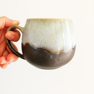 Puede incluir: Una taza de cerámica hecha a mano con un esmalte de dos tonos único. La mitad superior es de un verde claro y apagado, que pasa a una base marrón oscuro. La taza tiene una forma redondeada y un asa robusta.