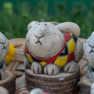 Op de afbeelding: Drie keramische konijnenfiguren met beschilderde details. Het konijn in het midden draagt een rood, geel en zwart gepatroneerd vest. Het konijn links draagt een blauw en wit gepatroneerd vest. Het konijn rechts draagt een blauw en wit gepatroneerd vest.