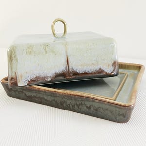 May include: A rectangular ceramic butter dish with a lid and a handle. The dish and lid are glazed in shades of blue, green, and brown. The lid has a small, round handle. The dish sits on a matching rectangular tray.