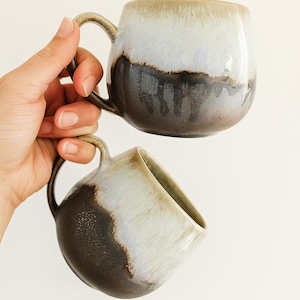 Op de afbeelding: Twee keramische mokken met een unieke glazuur. De mokken hebben een afgeronde vorm met een oor en een kleurovergang van lichtbeige naar donkerbruin. De mokken worden in een hand gehouden tegen een witte achtergrond. De mokken zijn geschikt voor koffie of thee.