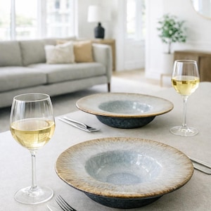 Könnte beinhalten: Zwei große, flache Keramikschalen mit blau-grauem Innenraum und braunem Rand stehen auf einer hellen Oberfläche. Zwei Weingläser mit Weißwein und Besteck stehen ebenfalls auf dem Tisch, was auf eine Essumgebung hindeutet.