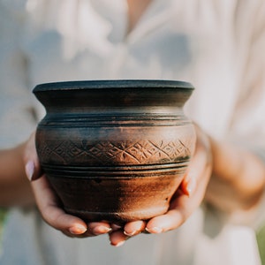 Puede incluir: Una olla de barro marrón con un borde marrón oscuro y tallas intrincadas. La olla está siendo sostenida por las manos de una persona.